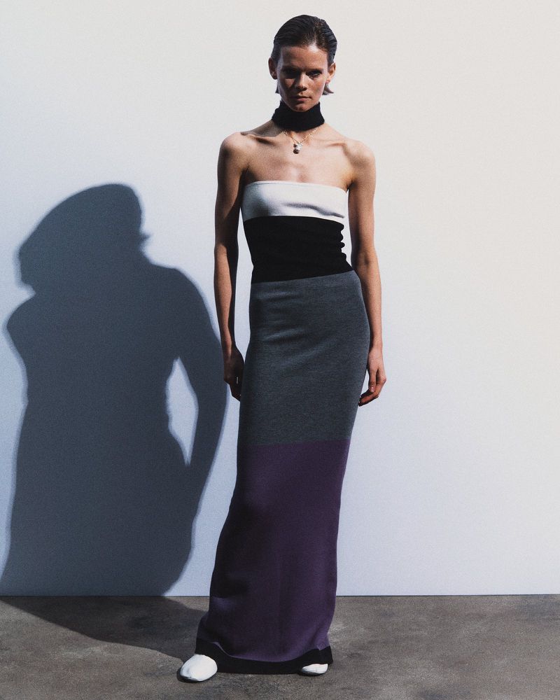 Fashion shot of a woman wearing a white, black, and grey tube top dress standing in front of a white wall with a shadow.