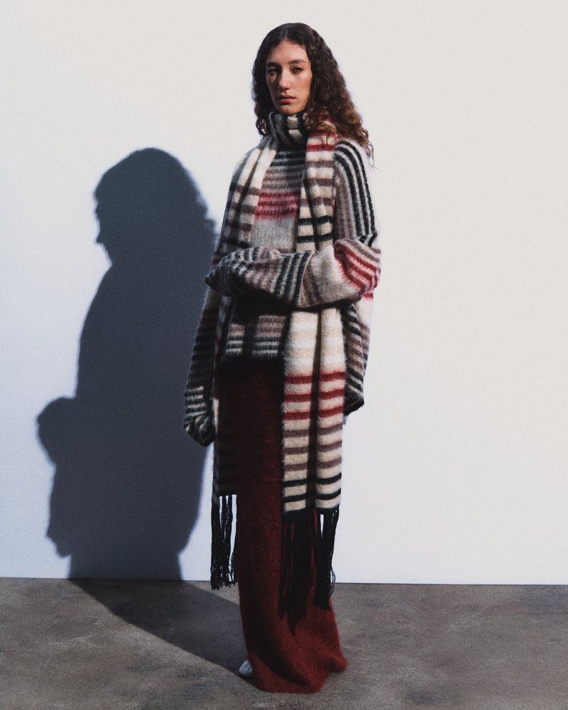 Model wearing a striped mohair knit sweater with matching fringe scarf and a long burgundy skirt against a studio wall.