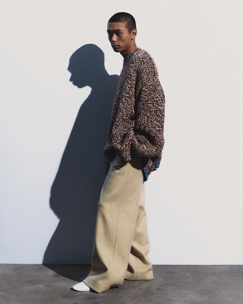 Asian man poses in studio wearing brown sweater and beige pants. Soft, diffused light, long shadow, fashion-forward editorial look.
