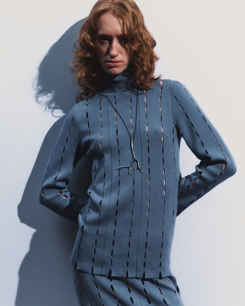 Adult female fashion model in blue knit turtleneck sweater and skirt with modern silver pendant necklace in a photo studio setting.