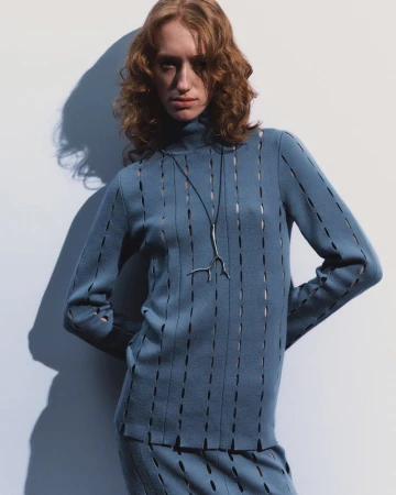 Adult female fashion model in blue knit turtleneck sweater and skirt with modern silver pendant necklace in a photo studio setting.