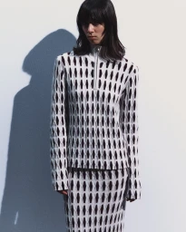 Model wearing a black and white geometric knit zip-up sweater and matching skirt against a white studio backdrop with a cast shadow.