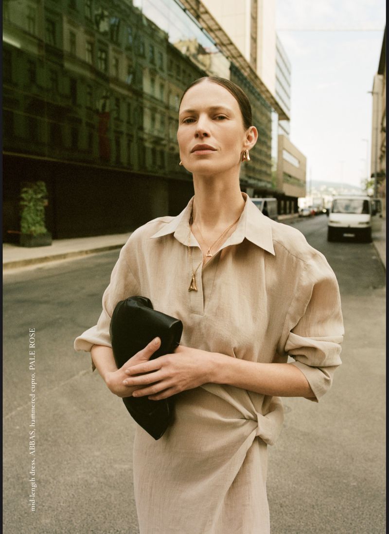 Fashion editorial shot of a woman in a beige linen dress with a black clutch purse on a city street with modern architecture.