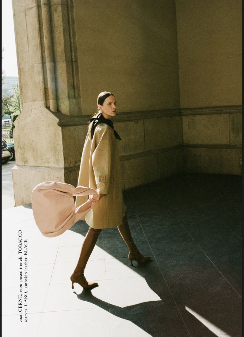 A fashionable adult female walks through an arched passageway holding a tan purse, wearing a trench coat, brown high heel boots and a black scarf.