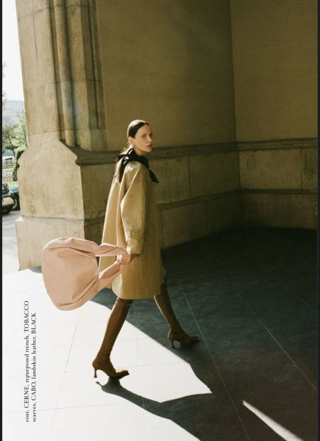 A fashionable adult female walks through an arched passageway holding a tan purse, wearing a trench coat, brown high heel boots and a black scarf.