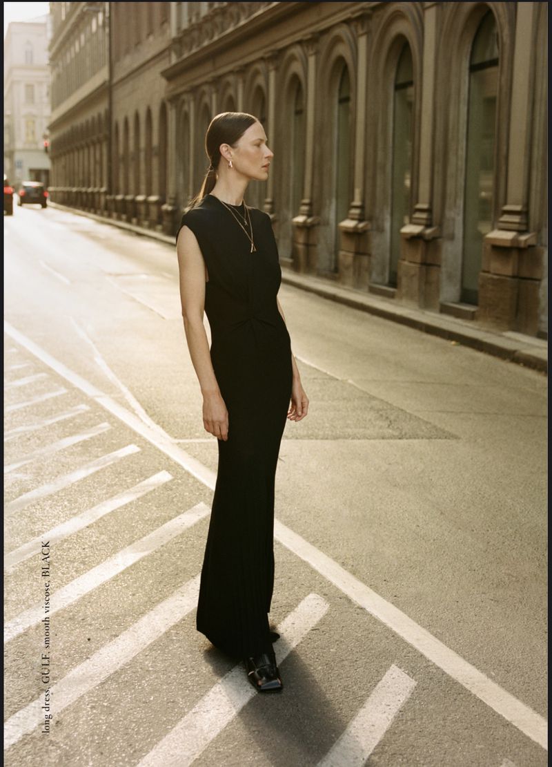 Woman in black dress stands on city street with light streaming on the road, tall buildings lining the street behind her.