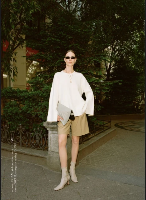 A woman in a cream sweater and beige shorts stands on a city street next to a building with trees, posing with a grey clutch bag.