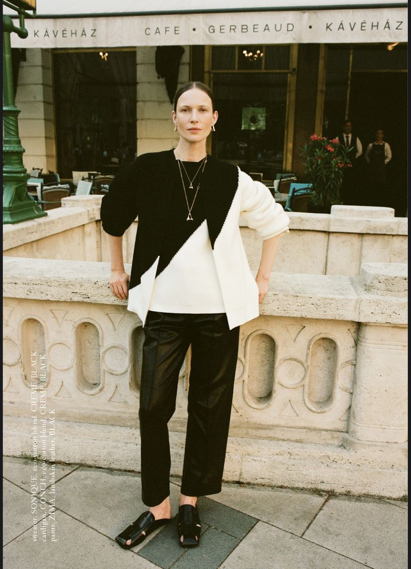 Woman in black and white sweater posing in front of Café Gerbeaud. She wears black pants and sandals, with natural light and urban setting.