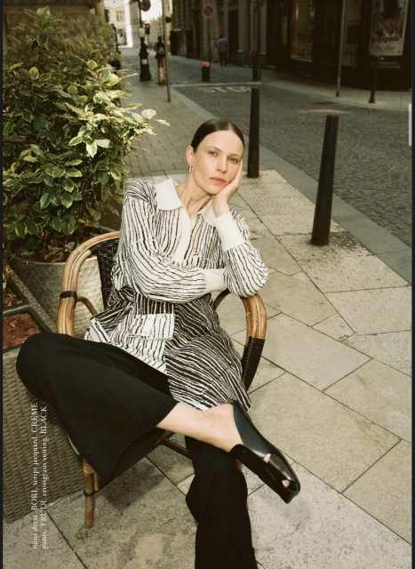 Adult female model sits on a rattan chair in an urban setting. She wears a striped shirt and black pants with a serious expression.