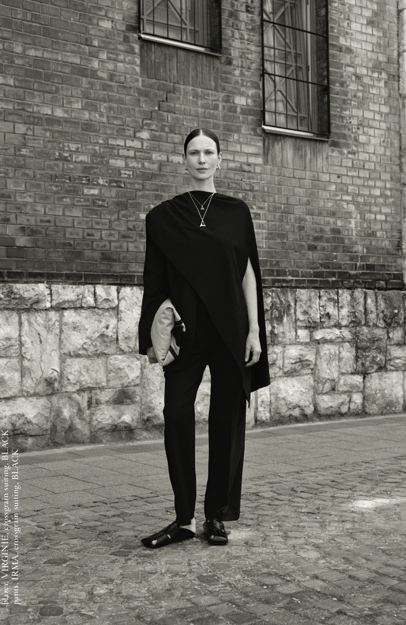 Monochrome portrait of a woman in draped black clothing standing in front of a brick building with windows, accessorized with a necklace.