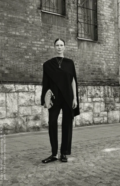 Monochrome portrait of a woman in draped black clothing standing in front of a brick building with windows, accessorized with a necklace.