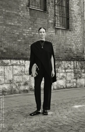 Monochrome portrait of a woman in draped black clothing standing in front of a brick building with windows, accessorized with a necklace.