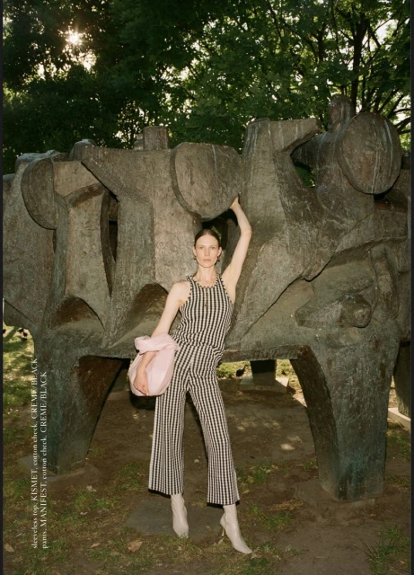 A tall woman stands in front of a large outdoor sculpture, wearing a striped jumpsuit and white boots, holding a pink bag, in a park setting.