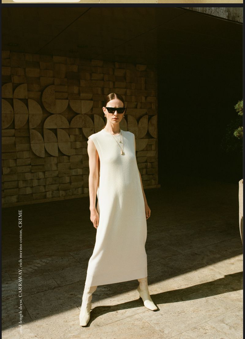 Woman poses in a long cream dress with sunglasses and boots, against a modern stone wall and dark archway, bathed in warm light.