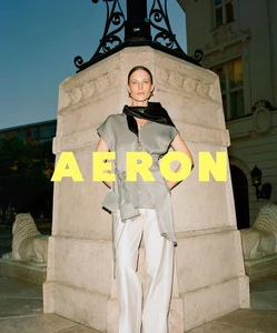 Model standing in front of a stone monument wearing a sage grey top and white trousers with yellow AERON text overlay