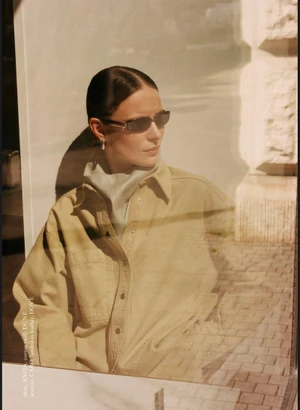Model Angelika Wierzbicka wearing narrow sunglasses and a sand-colored shirt behind reflective window glass.