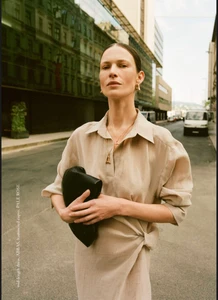 Angelika Wierzbicka posing outdoors in a draped beige collared dress while holding a black leather clutch on an urban street.
