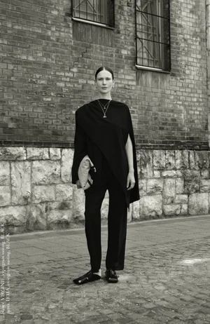 Model Angelika Wierzbicka posing outdoors in an asymmetrical black draped tunic, black trousers, and slides in front of a brick wall.