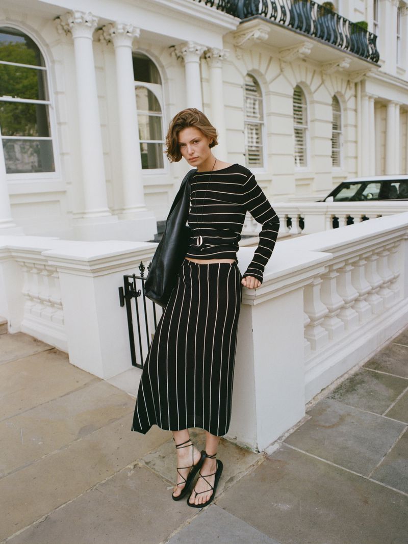 Adult woman in striped top and skirt poses on sidewalk with black leather jacket and strappy sandals in an urban environment.