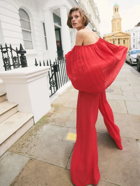 A woman in a red off-the-shoulder jumpsuit stands on a city street, looking over her shoulder toward the camera, posing for a fashion shot.