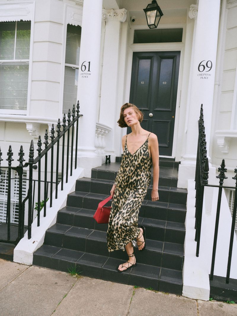 A woman in a leopard print dress descends the front steps of a London townhouse, holding a red bag with black stair railings.