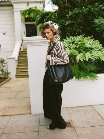 A stylish woman in a striped shirt and black pants poses outdoors with a black leather tote bag, near a building and lush greenery.