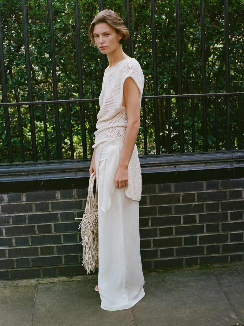 Adult woman in a cream-colored knit top and white pants stands outdoors with a macrame bag against a dark brick and wrought iron fence.