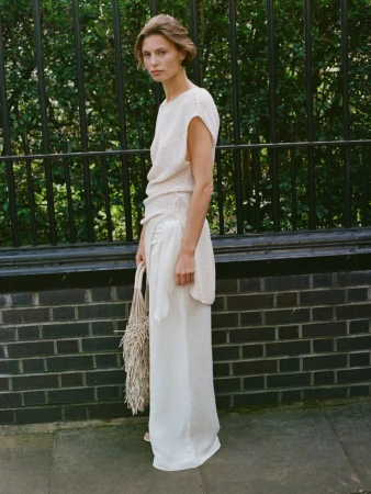 Adult woman in a cream-colored knit top and white pants stands outdoors with a macrame bag against a dark brick and wrought iron fence.