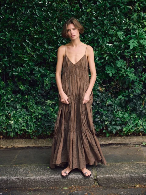 A woman with short hair stands against a green foliage background, wearing a flowing brown dress and sandals, captured in a medium shot.