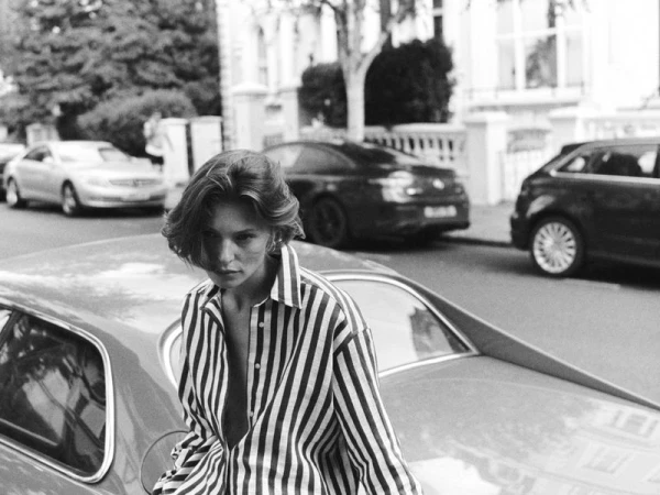 Monochrome portrait of a young woman in a striped shirt, posing near a car on a street with parked vehicles and buildings.