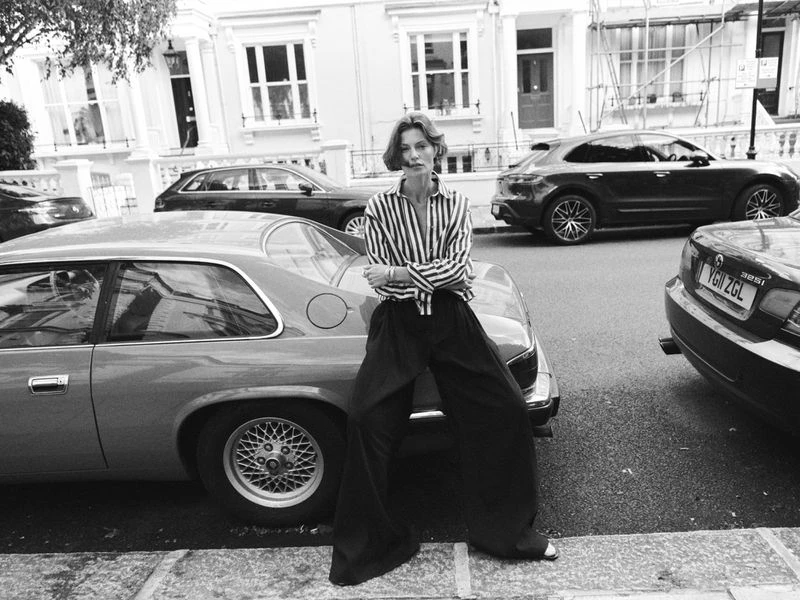 Black and white shot of an adult woman with short hair wearing a striped shirt and dark pants, leaning against a vintage car in a city street.