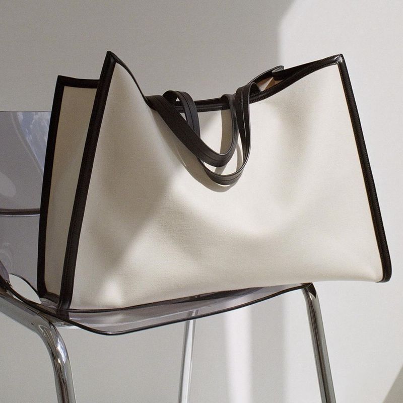 A large, off-white tote bag with dark brown trim sits on a clear acrylic chair against an off-white wall in a neutral-toned room.