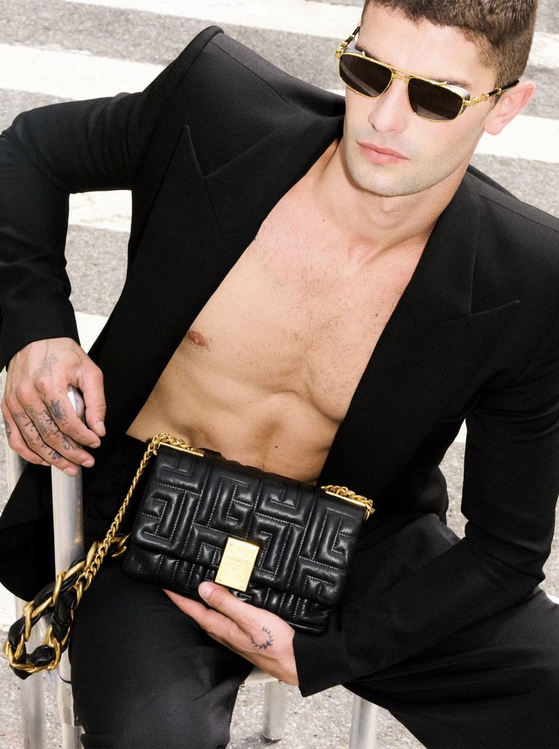 Stylish man in a black blazer and trousers holds a black quilted purse with a gold chain strap while sitting on metal scaffolding.