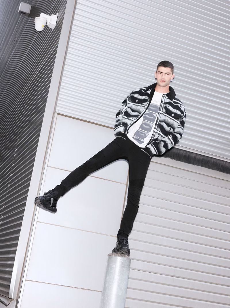 Male model strikes a pose in black and white zebra-print jacket and black jeans against a corrugated metal wall outdoors during daytime.