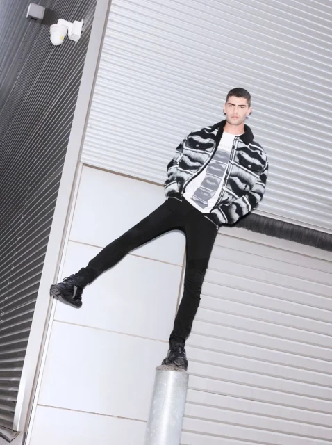 Male model strikes a pose in black and white zebra-print jacket and black jeans against a corrugated metal wall outdoors during daytime.