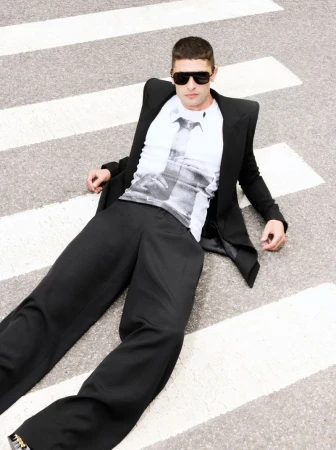 Man wearing a black blazer, white graphic t-shirt, and black trousers lying on a pedestrian crosswalk.