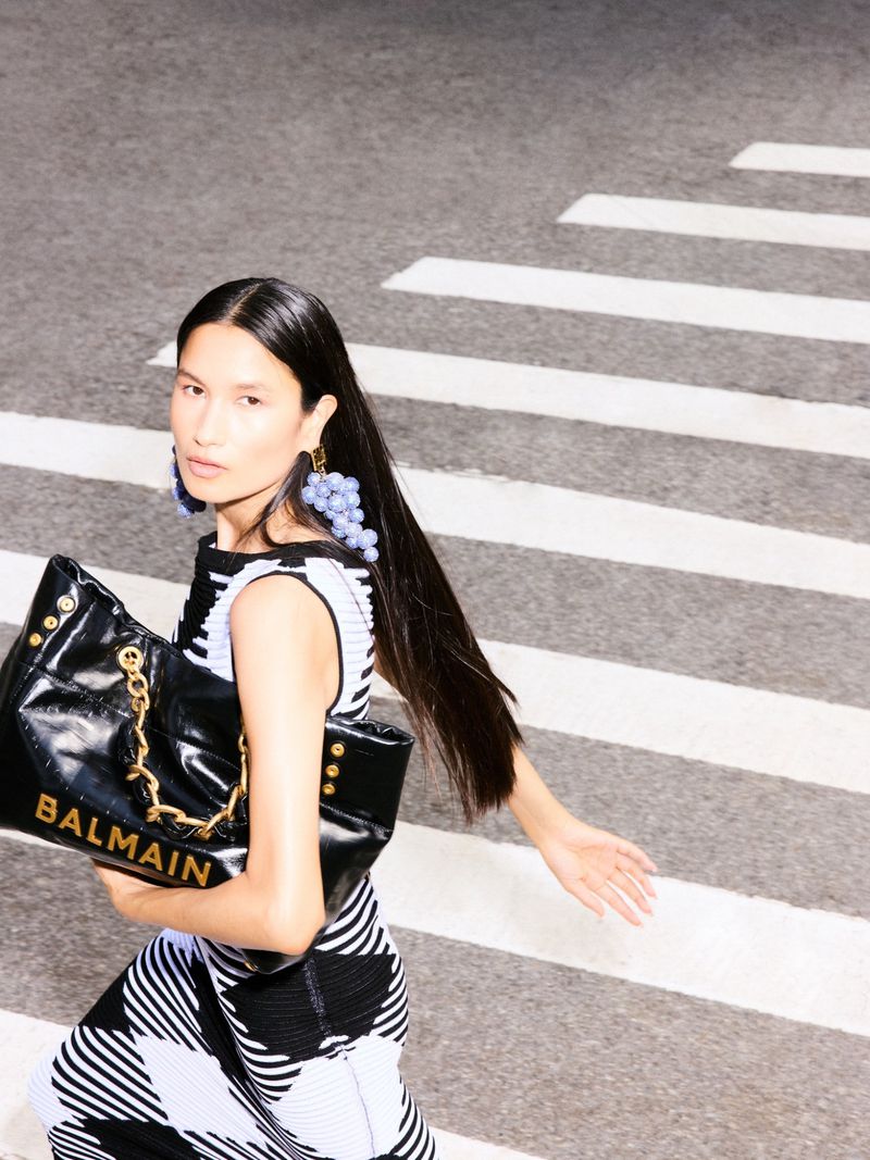 Adult female in black-and-white dress with Balmain purse on crosswalk with grape earrings in fashion editorial shot.