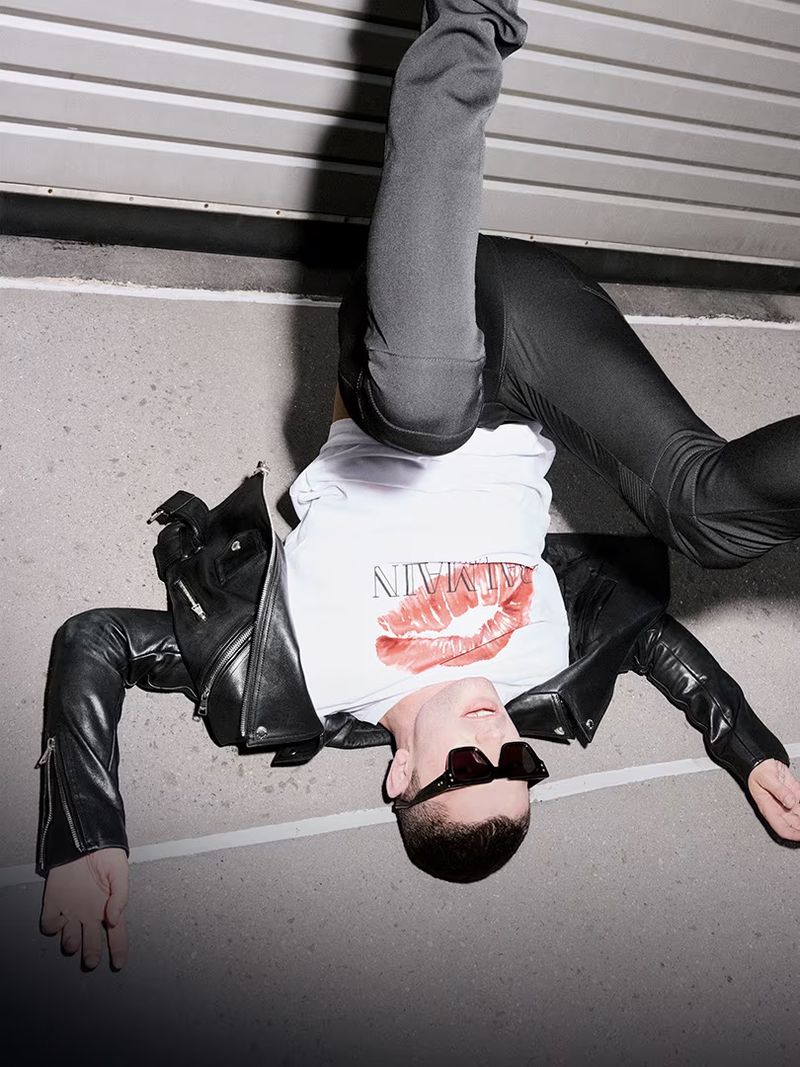 Upside-down fashion shot of a man wearing a branded t-shirt, black jacket, sunglasses, and leggings lying on a concrete surface.