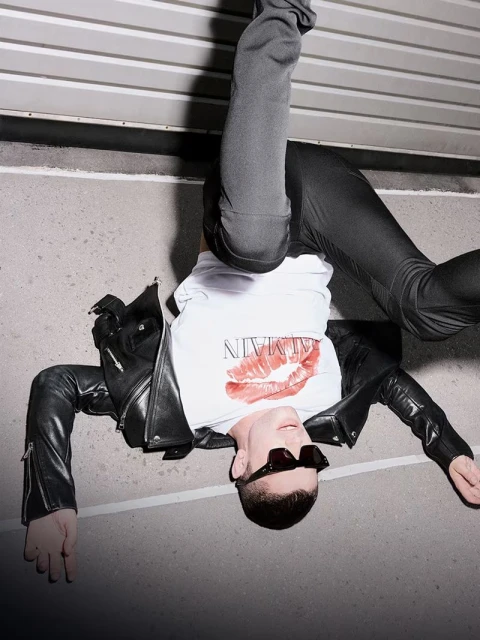 Upside-down fashion shot of a man wearing a branded t-shirt, black jacket, sunglasses, and leggings lying on a concrete surface.