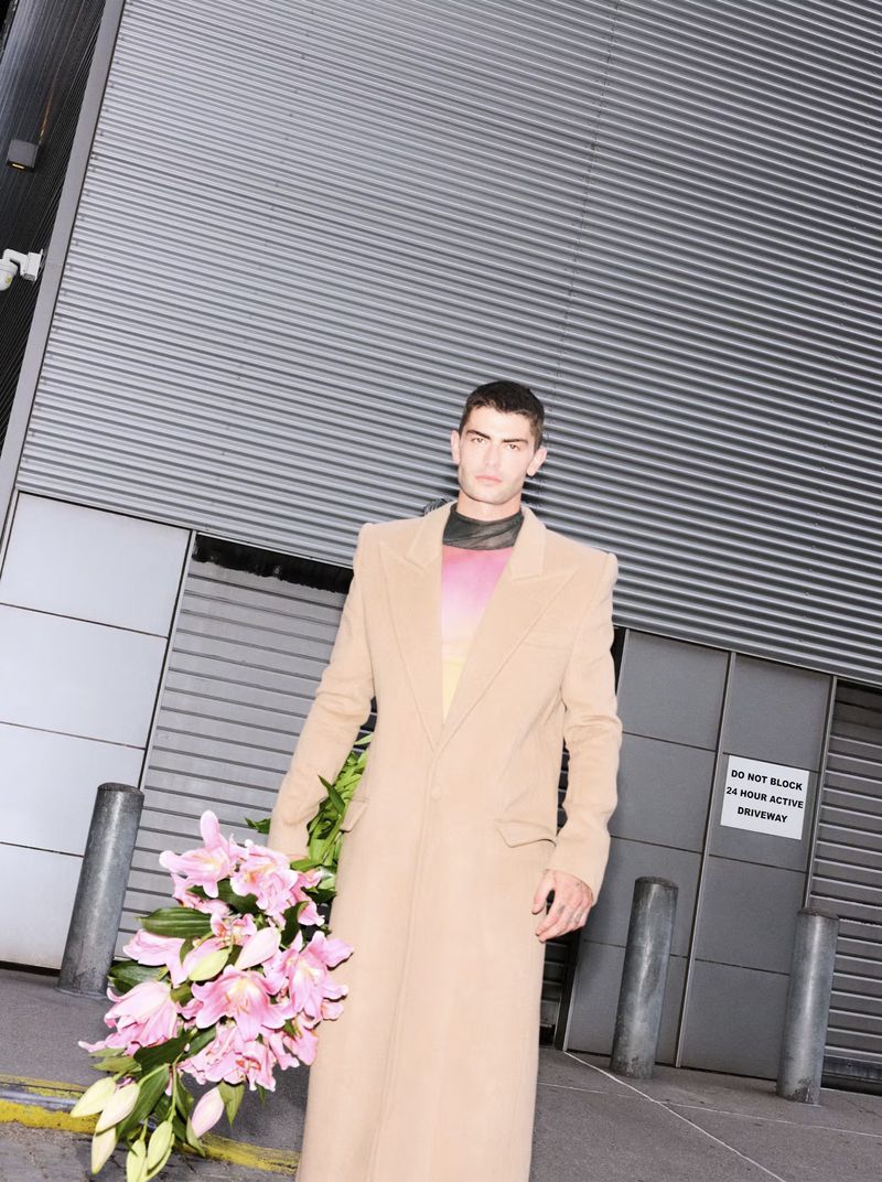 A stylish adult male with a tan overcoat and a bouquet of pink lilies stands outside an industrial building with grey corrugated metal.