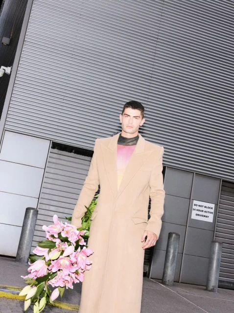 A stylish adult male with a tan overcoat and a bouquet of pink lilies stands outside an industrial building with grey corrugated metal.