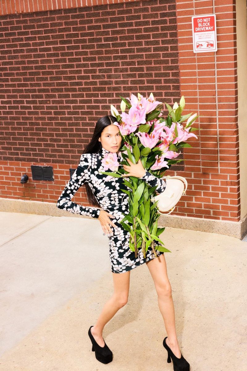 A fashionable adult woman poses outdoors on pavement holding a large bouquet of pink lilies and an off-white purse with a brick wall backdrop.
