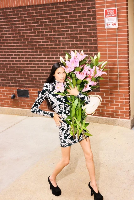 A fashionable adult woman poses outdoors on pavement holding a large bouquet of pink lilies and an off-white purse with a brick wall backdrop.