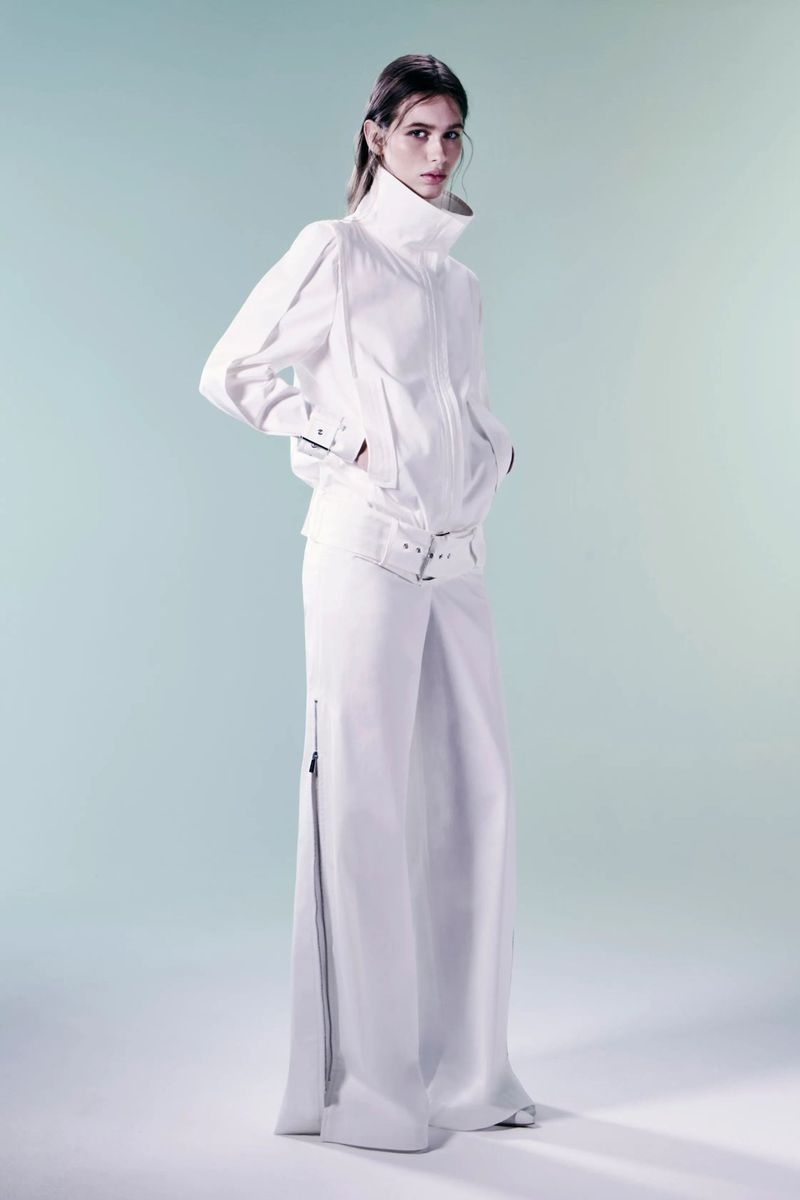 Fashion photo of a young adult female model wearing a high-collar white jacket and wide-leg pants with zippers against a light blue background.