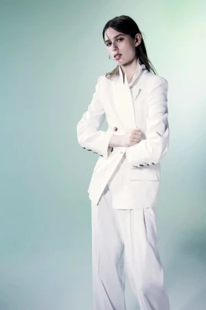Fashion portrait of a young woman in an all-white double-breasted suit. Studio shot against a gradient light-blue background.