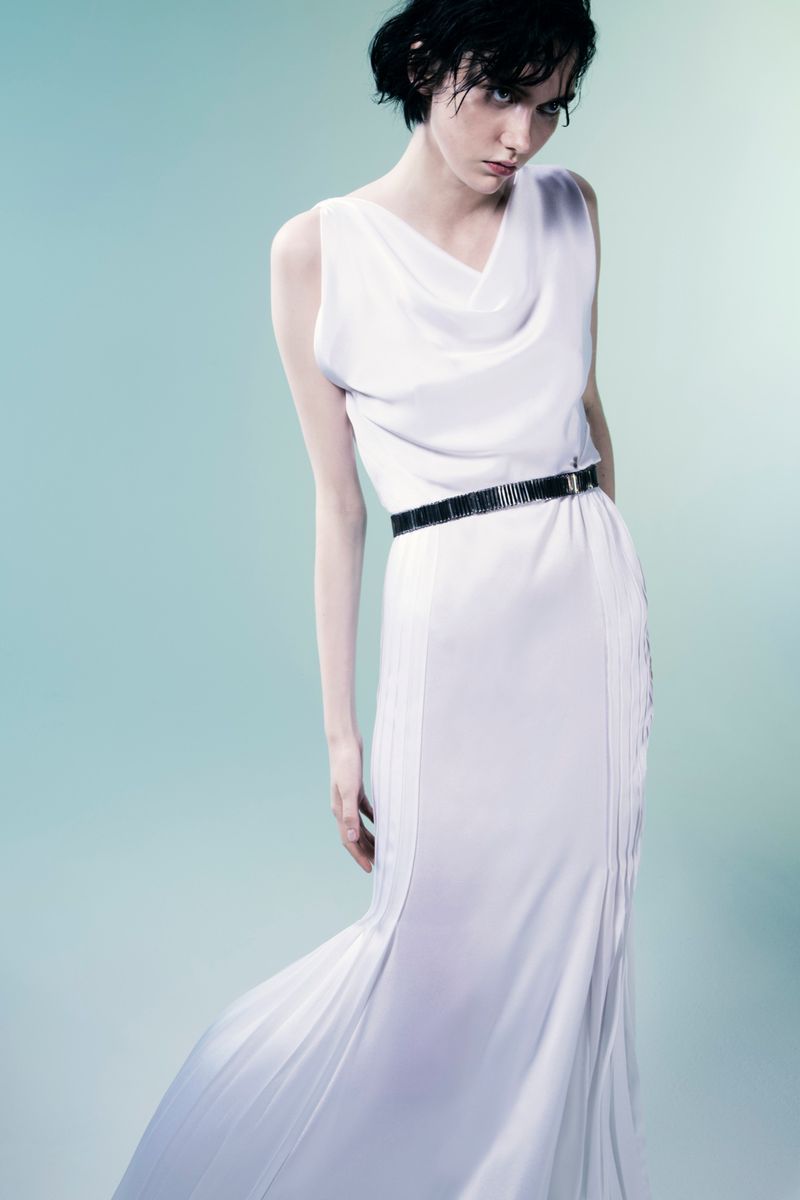 Model posing in a draped sleeveless white floor-length gown with a thin black belt against a pale cyan gradient studio backdrop.