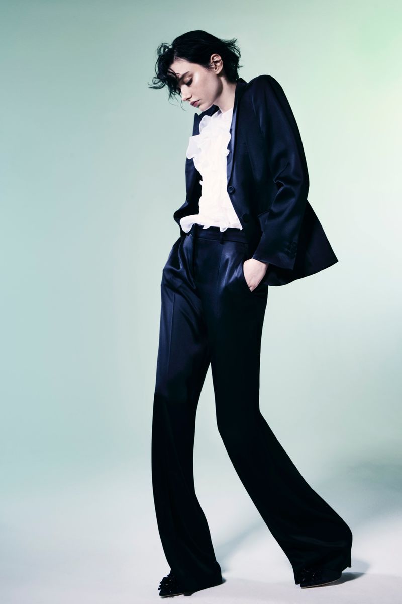 A woman poses in a dark blue silk suit with a white ruffled blouse against a soft aqua studio background, fashion-forward style.