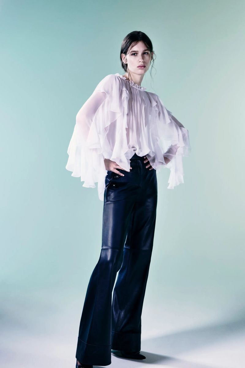 A slender woman poses indoors, wearing a flowing white blouse and shiny black flared pants, illuminated by bright lighting.