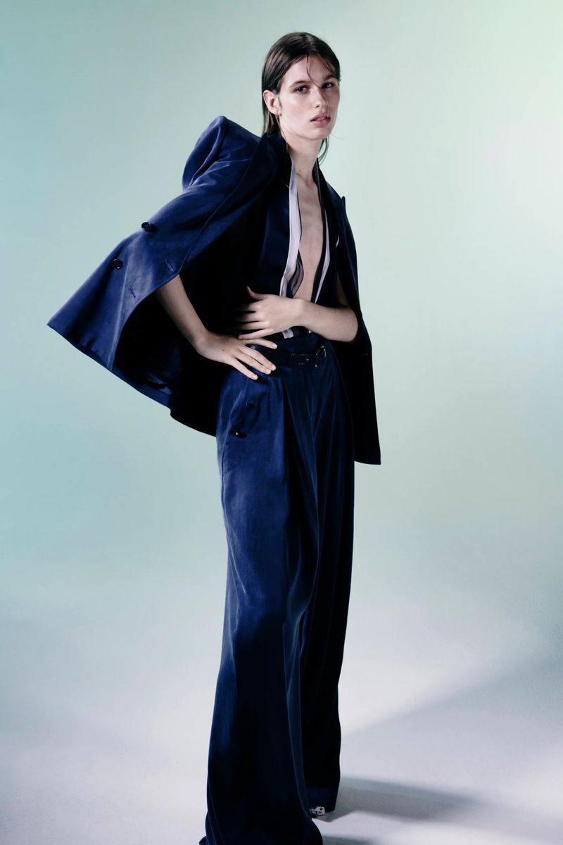 A fashion photo showcases a woman in a navy suit with an open jacket, wide-leg pants, against a light blue backdrop in a studio setting.