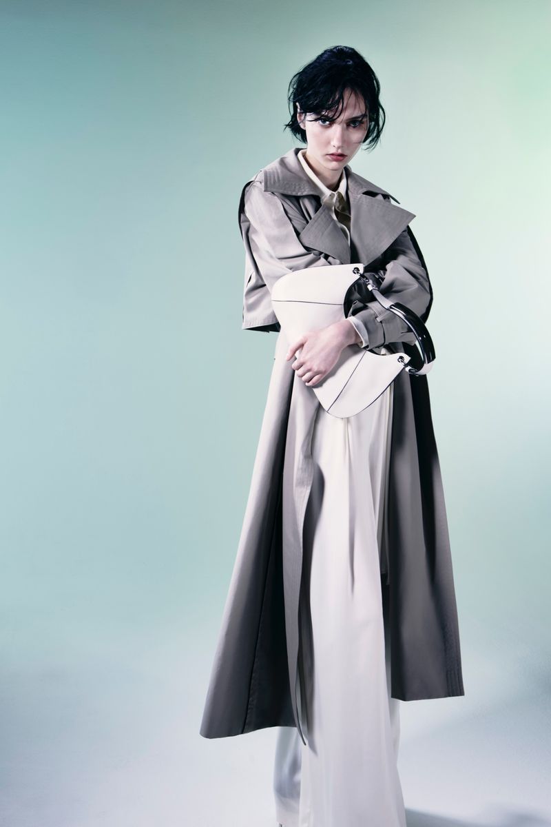 Model Steinberg wearing an oversized beige layered trench coat and clutching a white curved handbag against a pale studio backdrop.